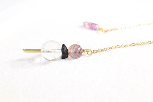 side view of pendulum, the weight has a brass pointer, a large clear quartz bead, then a black tourmaline chip, and it topped with a fluorite bead, it has a dainty gold chain and on the other end is a rectangular fluorite holder. pendulum is laying on white sand, and photo is taken from the side on a low angle.