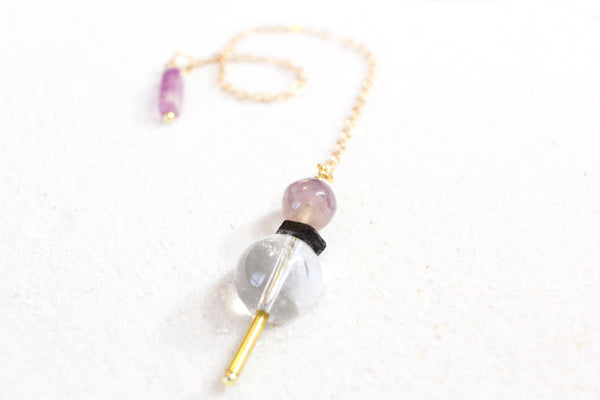 front view of pendulum, the weight has a brass pointer, a large clear quartz bead, then a black tourmaline chip, and it topped with a fluorite bead, it has a dainty gold chain and on the other end is a rectangular fluorite holder. pendulum is laying on white sand, and photo is taken from the front on a low angle.