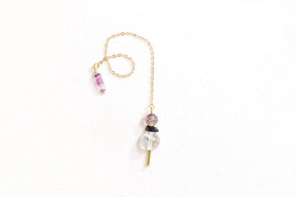 top view of pendulum, the weight has a brass pointer, a large clear quartz bead, then a black tourmaline chip, and it topped with a fluorite bead, it has a dainty gold chain and on the other end is a rectangular fluorite holder. pendulum is laying on white sand