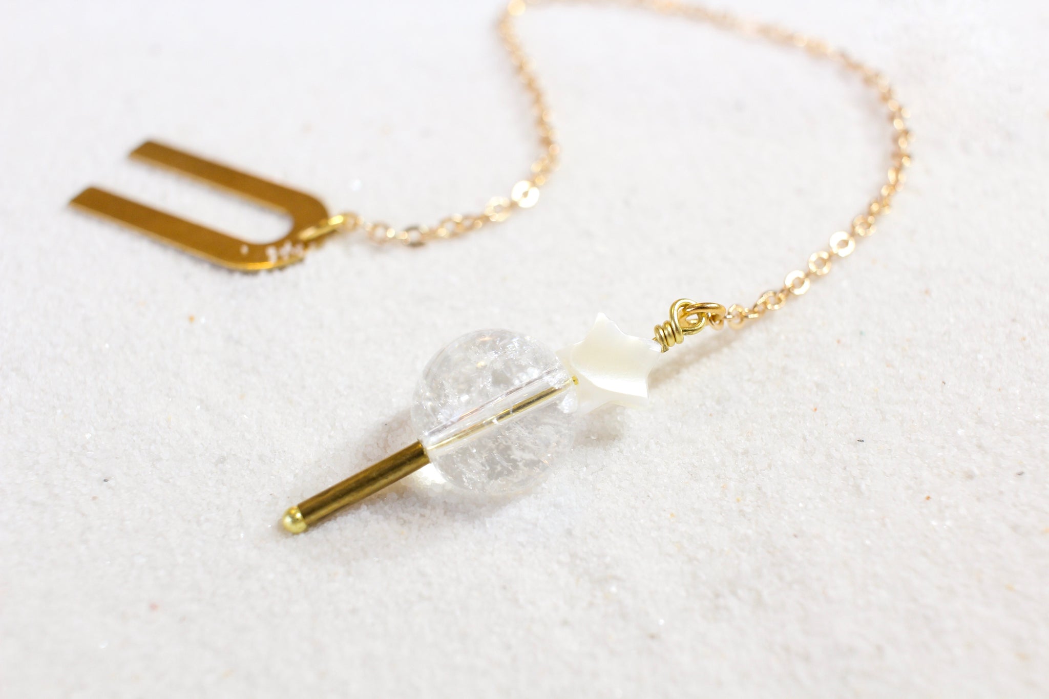 Pendulum with a gold chain and a gold arch shaped holder. the end is a clear quartz weight, with a brass pointer underneath and a white mother of pearl star on top. laying on white sand.