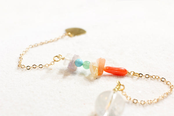 white sand with a delicate gold chain pendulum. the pendulum has chakra-themed beads on the chain, and a clear quartz end with little gold beads for a pointer at the end. close up of the chakra beads, which are white kunzite, amethyst, turquoise howlite, aventurine, citrine, sunstone + red coral