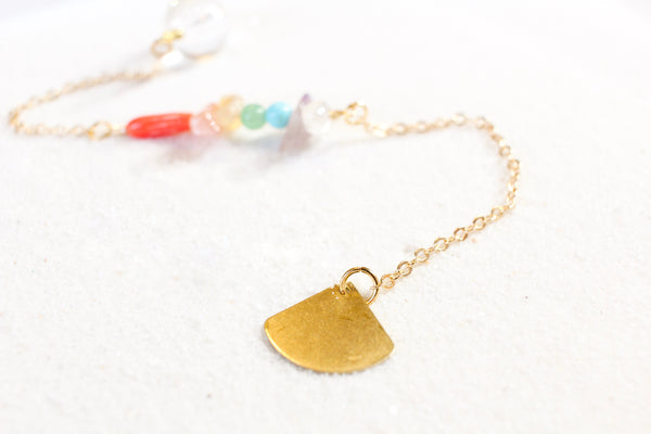 white sand with a delicate gold chain pendulum. the pendulum has chakra-themed beads on the chain, and a clear quartz end with little gold beads for a pointer at the end. zoom in of the fan-shaped gold brass hold end