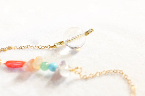 white sand with a delicate gold chain pendulum. the pendulum has chakra-themed beads on the chain, and a clear quartz end with little gold beads for a pointer at the end.