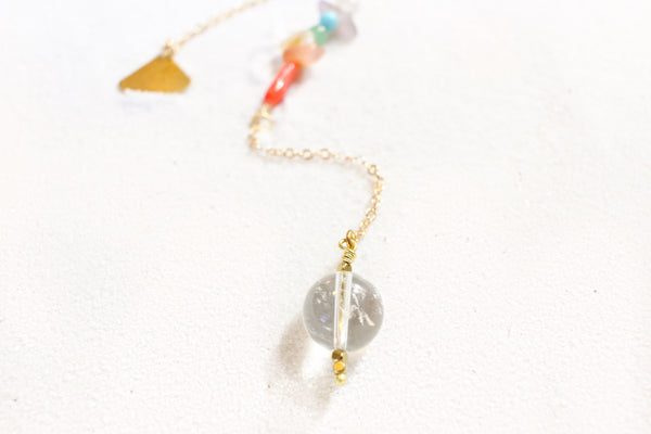 white sand with a delicate gold chain pendulum. the pendulum has chakra-themed beads on the chain, and a clear quartz end with little gold beads for a pointer at the end. close up of the clear quartz pendulum end
