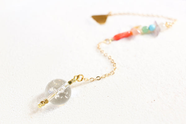 white sand with a delicate gold chain pendulum. the pendulum has chakra-themed beads on the chain, and a clear quartz end with little gold beads for a pointer at the end.