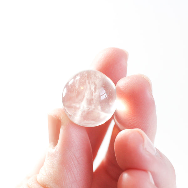 .75 inch clear quartz sphere being held in the fingers of a caucasian woman- the light is shining through the quartz in a warm, angelic way