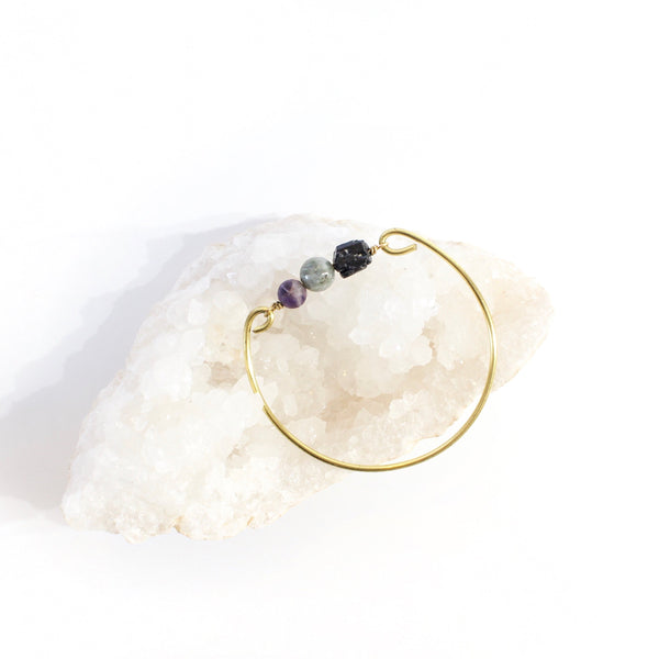 white background with a gold brass bangle. on the bangle is raw back tourmaline, a labradorite sphere bead and an amethyst sphere bead