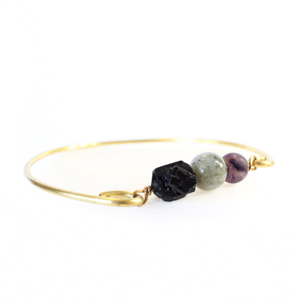 white background with a gold brass bangle. on the bangle is raw back tourmaline, a labradorite sphere bead and an amethyst sphere bead. 45 degree angle shot.