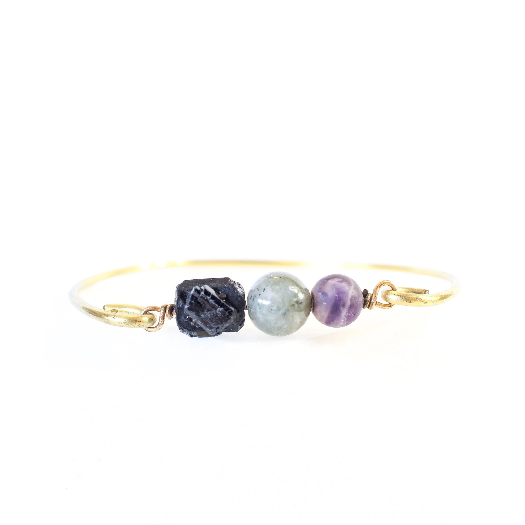 white background with a gold brass bangle. on the bangle is raw back tourmaline, a labradorite sphere bead and an amethyst sphere bead