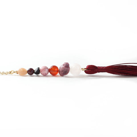 String of beads, with dainty gold chain loop on one end, and a dark red tassel on the other end. The beads are as follows (from top to bottom): sunstone, garnet, black tourmaline, rhodonite, carnelian, pink tourmaline and rose quartz. Tassel is laying on it's side with a completely white background.