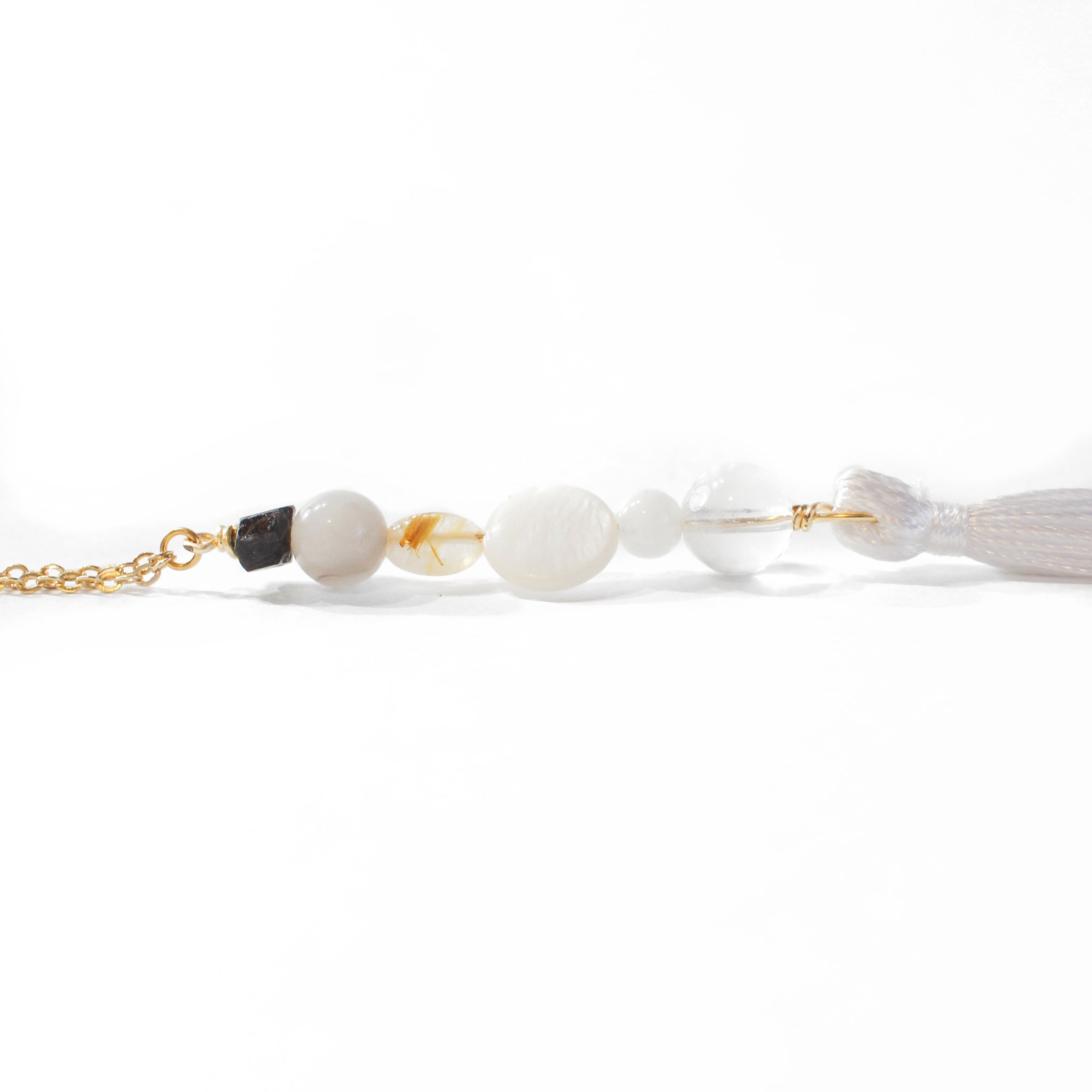 String of beads, with a delicate gold chain on one end and a white tassel on the other. The string of beads (from top to bottom) is black tourmaline, grey agate, gold rutilated quartz, mother of pearl, moonstone, and clear quartz. Tassel is laying on it's side with a completely white background