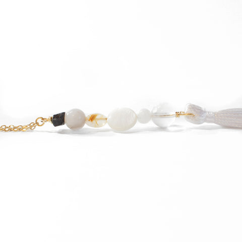 String of beads, with a delicate gold chain on one end and a white tassel on the other. The string of beads (from top to bottom) is black tourmaline, grey agate, gold rutilated quartz, mother of pearl, moonstone, and clear quartz. Tassel is laying on it's side with a completely white background