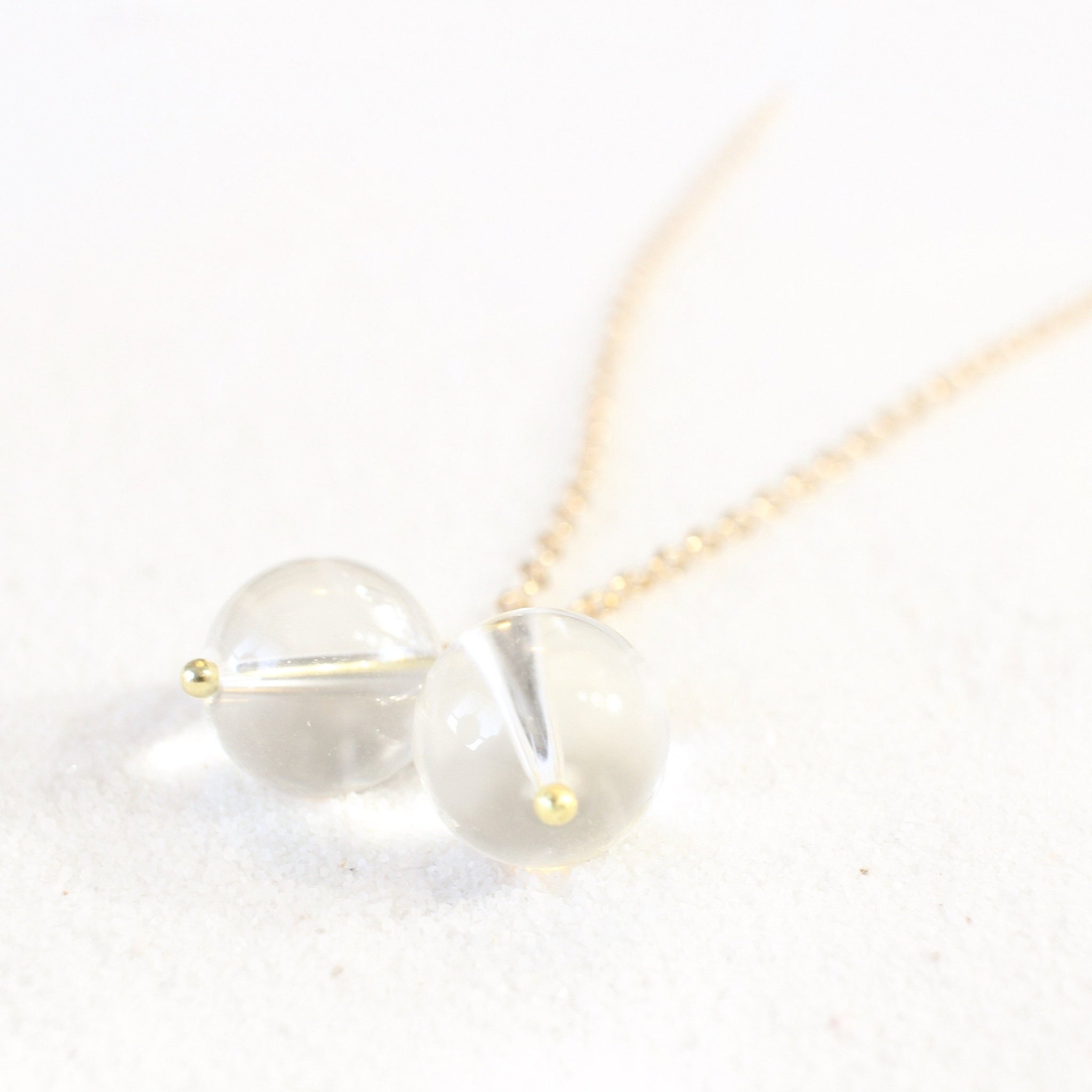 dainty gold chain with two quartz pendants that look like round breasts with gold nipples., laying on white sand, photo is a view from the bottom of the pendant