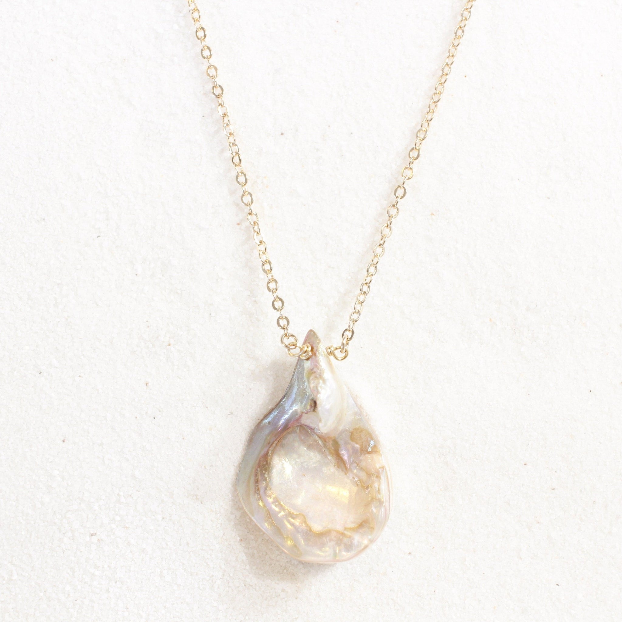 iridescent seashell pendant with a striking resemblance to a vulva, on a dainty gold chain. necklace is laying on white sand.