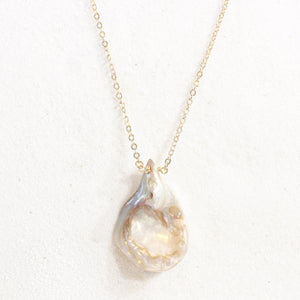 iridescent seashell pendant with a striking resemblance to a vulva, on a dainty gold chain. necklace is laying on white sand.