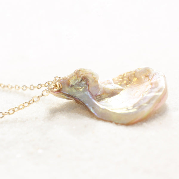 iridescent seashell pendant with a striking resemblance to a vulva, on a dainty gold chain. necklace is laying on white sand. side view.