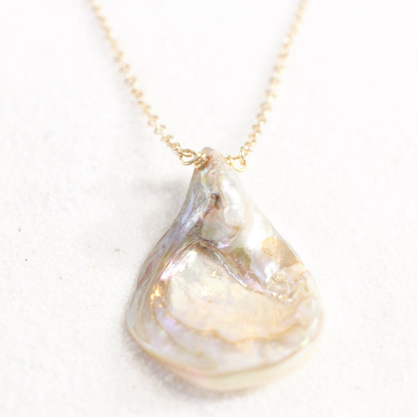 iridescent seashell pendant with a striking resemblance to a vulva, on a dainty gold chain. necklace is laying on white sand. view from the front, bottom of the pendant