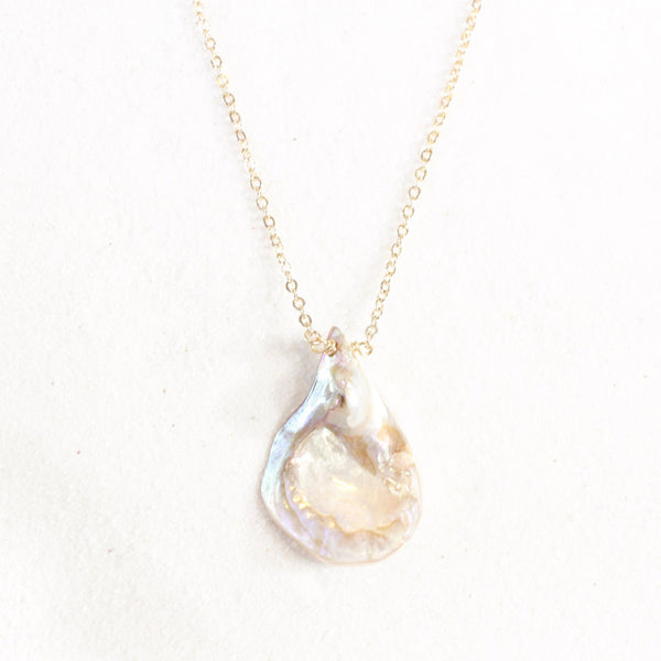 iridescent seashell pendant with a striking resemblance to a vulva, on a dainty gold chain. necklace is laying on white sand.