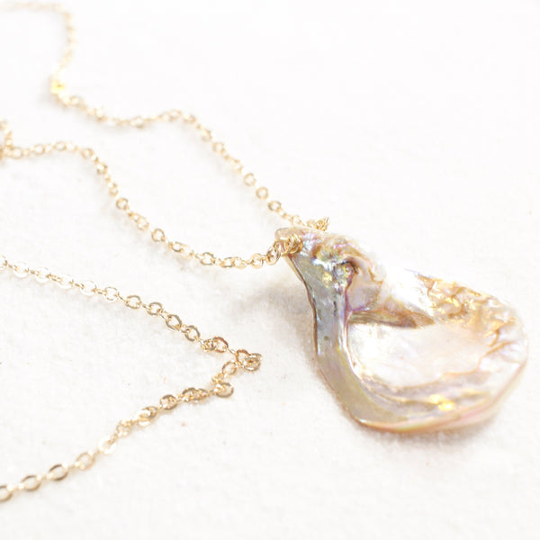 iridescent seashell pendant with a striking resemblance to a vulva, on a dainty gold chain. necklace is laying on white sand. 45 degree angle view