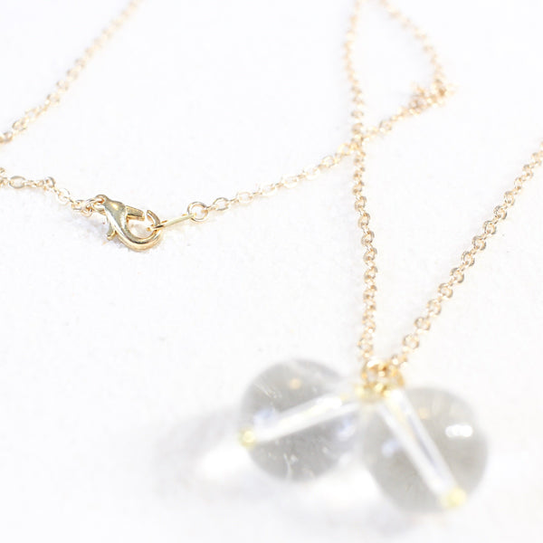 dainty gold chain with two quartz pendants that look like round breasts with gold nipples., laying on white sand, photo is a view from the bottom of the pendant, but is showcasing a clear image of a gold lobster clasp