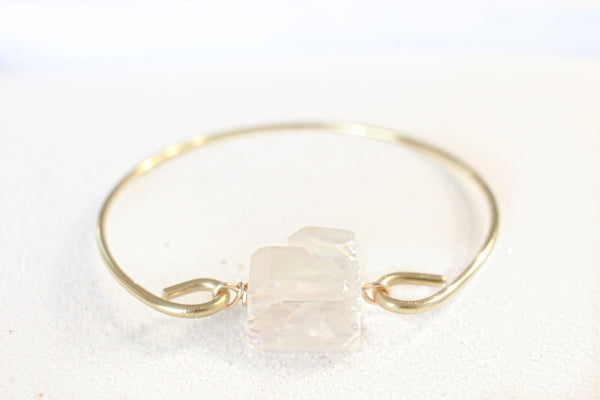 gold bangle with an aura quartz chunk on it, photo taken straight-on with the crystal, bracelet is shown laying on white sand