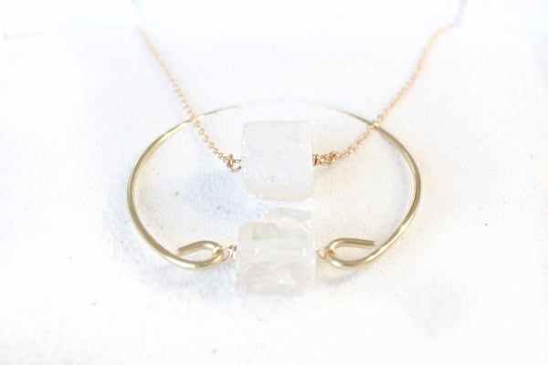 gold bangle with an aura quartz (clear quartz with iridescent coating) chunk on it. laying with a dainty gold necklace with aura quartz on it. they are laying on white sand