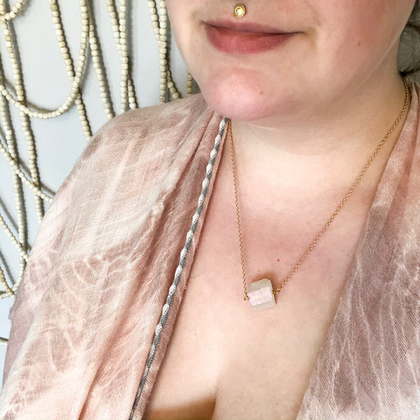 gold necklace with an aura quartz (clear quartz with an iridescent coating) chunk on it. necklace is mid chest length. necklace is shown worn by a caucasian woman. she is wearing a pink shawl.