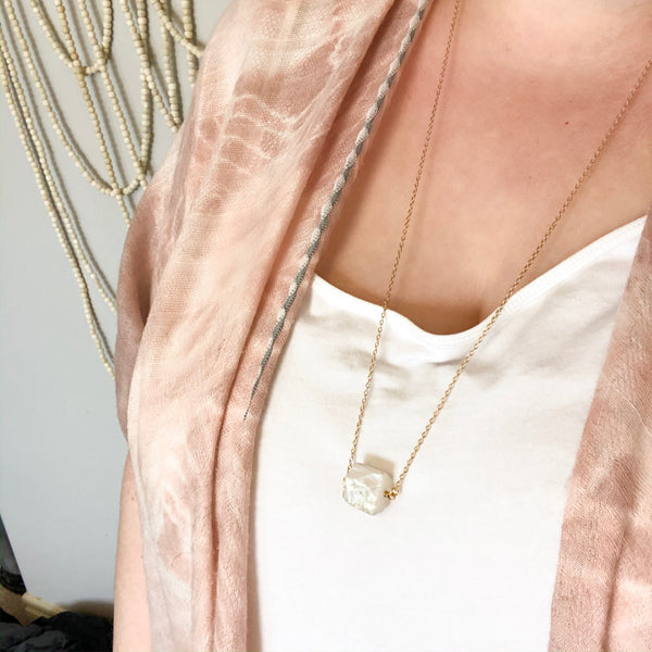 gold necklace with an aura quartz (clear quartz with an iridescent coating) chunk on it. necklace is long, mid breast length. necklace is shown worn by a caucasian woman. she is wearing a pink shawl.