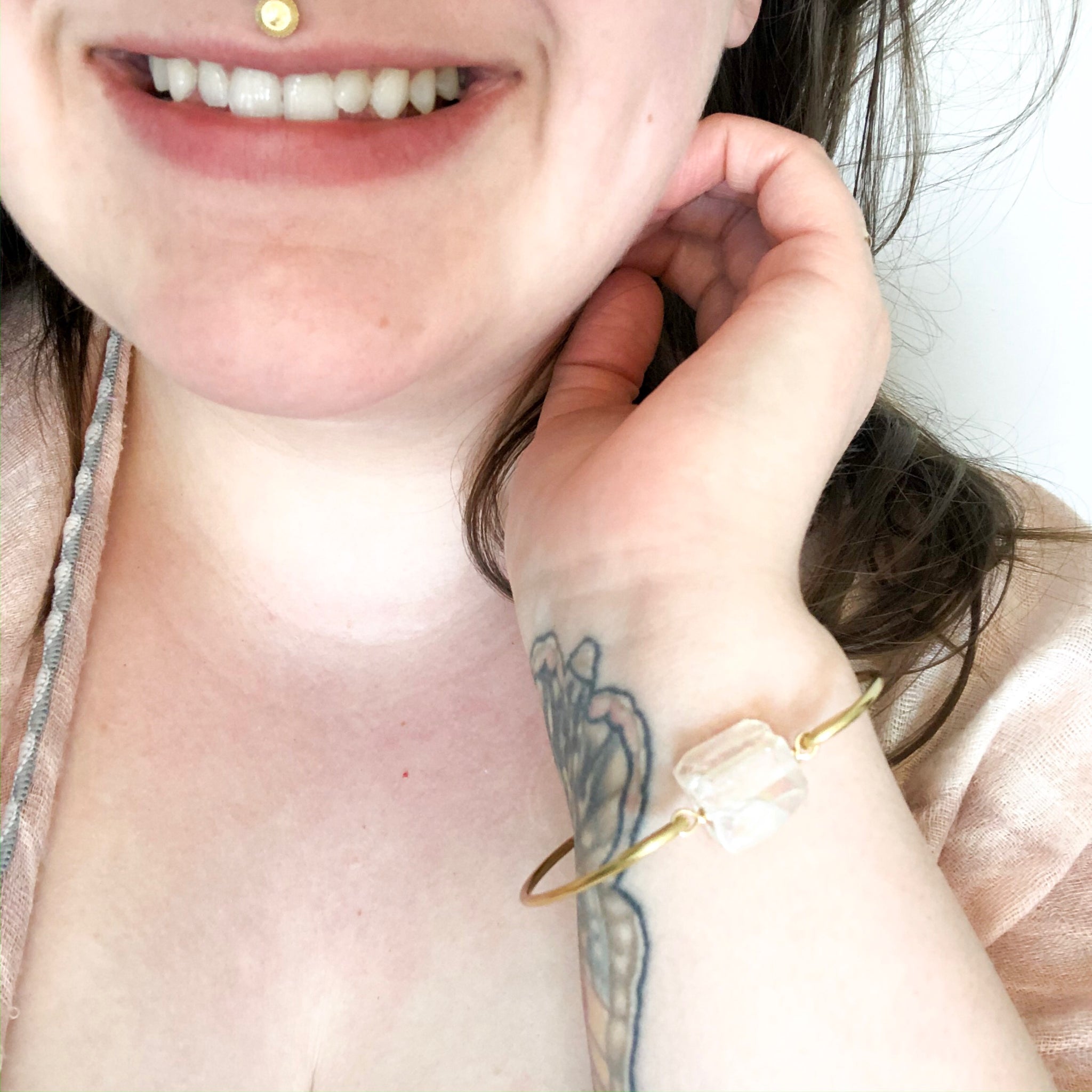 gold bangle with a aura quartz (clear quartz with an iridescent coating) chunk in the middle. bracelet is shown worn by a caucasian woman with brown hair. she is wearing a pink shawl and is smiling.