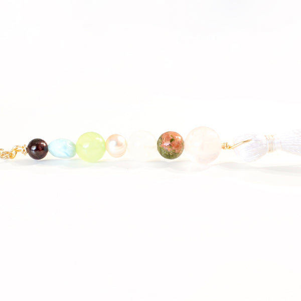 string of crystals with a loop of thin gold chain to hang it. the crystals from top to bottom are garnet, larimar, jade, pearl, moonstone, unakite and rose quartz. at the bottom to the crystals is a white tassel. white background.