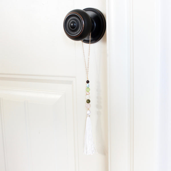 string of crystals with a loop of thin gold chain to hang it. the crystals from top to bottom are garnet, larimar, jade, pearl, moonstone, unakite and rose quartz. at the bottom to the crystals is a white tassel. door tassel is hanging on a black doorknob on a white door.