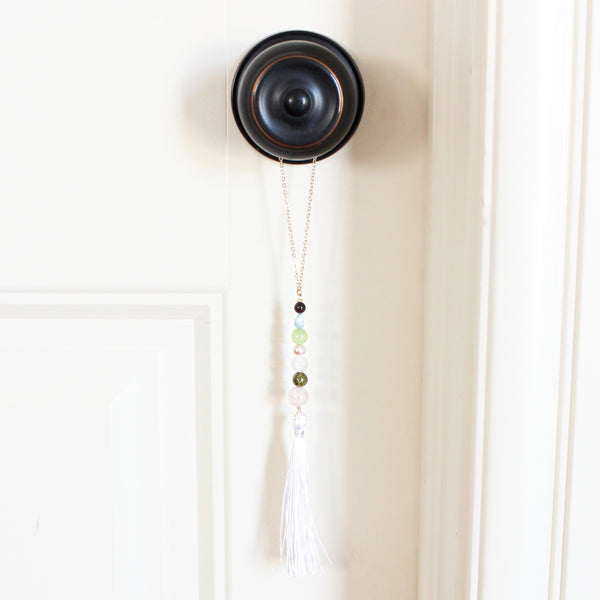 string of crystals with a loop of thin gold chain to hang it. the crystals from top to bottom are garnet, larimar, jade, pearl, moonstone, unakite and rose quartz. at the bottom to the crystals is a white tassel. door tassel is hanging on a black doorknob on a white door.