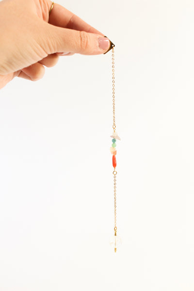 woman's hand holding the hold end of a pendulum with a delicate gold chain. the pendulum has chakra-themed beads on the chain, and a clear quartz end with little gold beads for a pointer at the end.