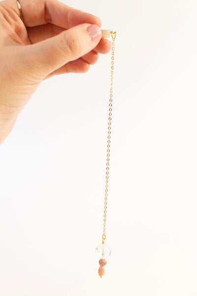 pendulum being held by holder, hanging in front of a white background. it has a lithium quartz holder, a gold chain, and a clear quartz, rhodonite and sunstone weight on the other end.