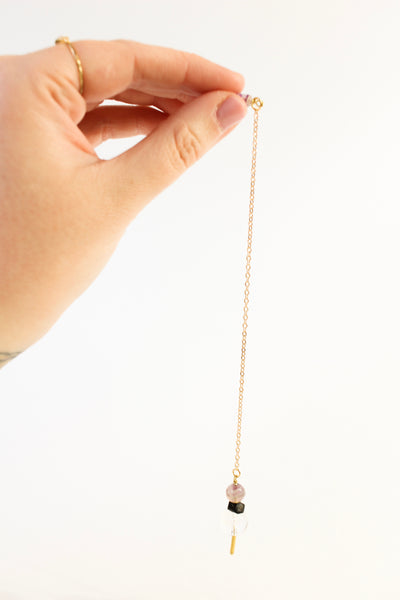 side view of pendulum, the weight has a brass pointer, a large clear quartz bead, then a black tourmaline chip, and it topped with a fluorite bead, it has a dainty gold chain and on the other end is a rectangular fluorite holder. pendulum is being held by a white woman's hand, hanging in front of a white background.