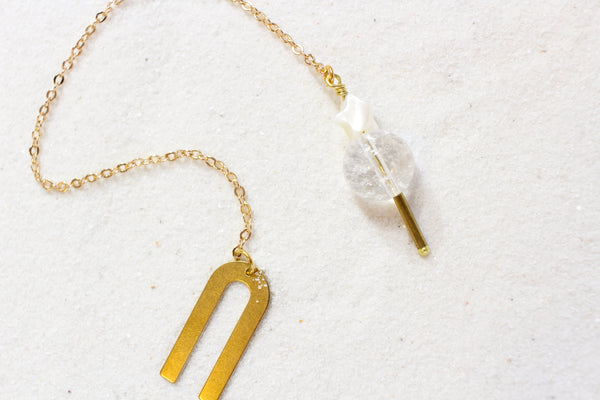 Pendulum with a gold chain and a gold arch shaped holder. the end is a clear quartz weight, with a brass pointer underneath and a white mother of pearl star on top. top view,  laying on white sand.