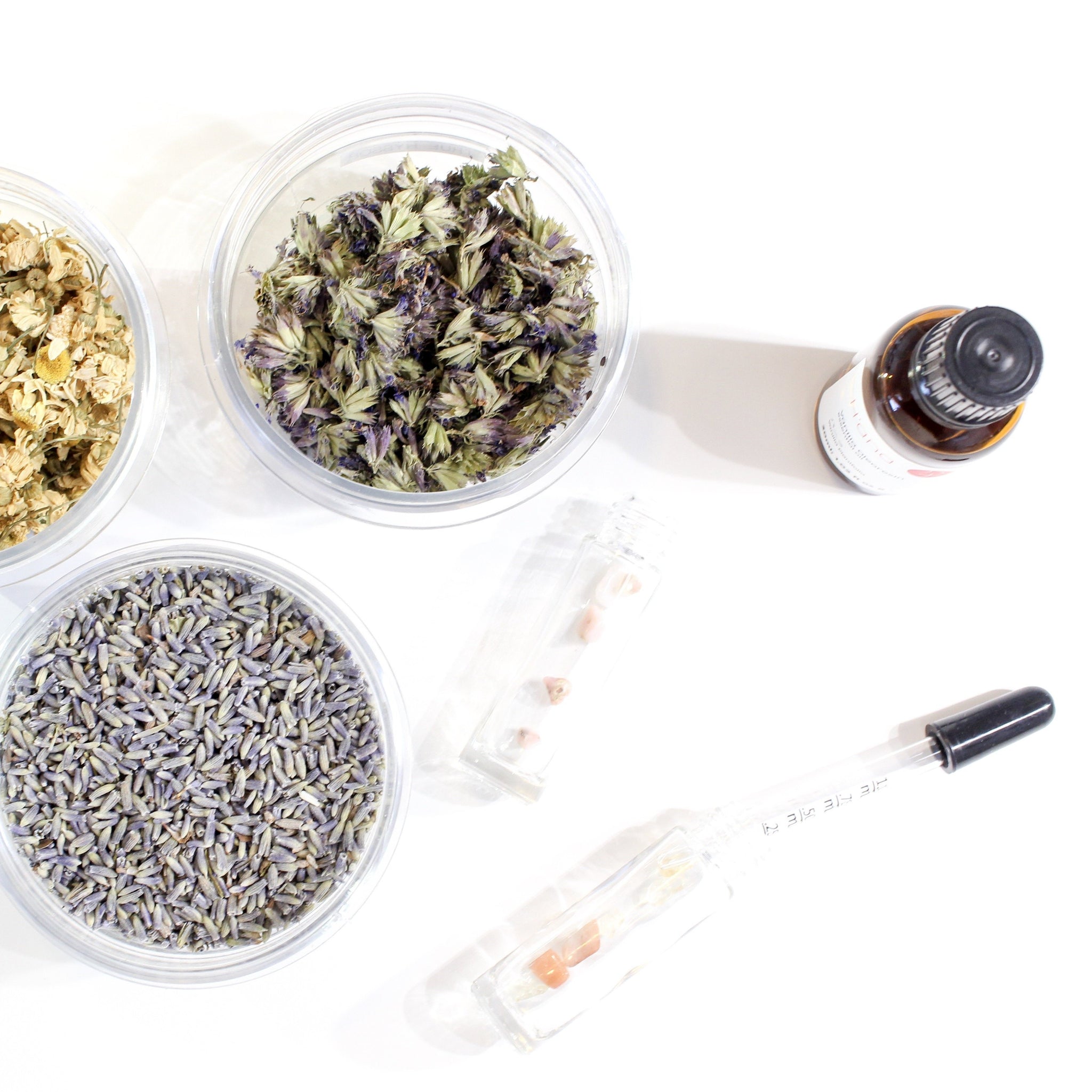 white background with three clear containers containing dried herbs. there is two empty clear roller bottles laying on their sides with crystals in them. one little pipette and one bottle of essential oil