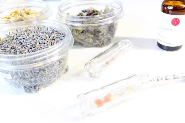 white background with clear containers holding dried herbs. there and clear glass roller bottles with crystals in them laying on their side, a glass pipette and a bottle of essential oil