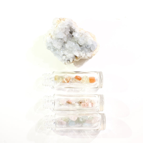 White background with three glass roller bottles filled with crystals laying on their sides, one on top of the other. on the very top is a soft blue crystal  cluster