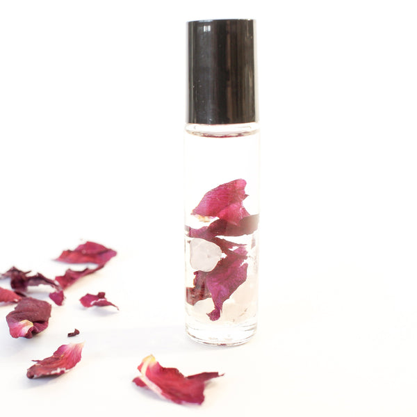 white background- clear glass roller bottle with a black lid. inside the bottle is rose quartz and dried red rose petals. there is a little trail of dried rose petals on the white surface that the bottle is standing on