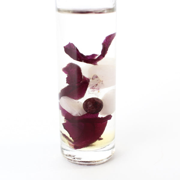 clear glass roller bottle with a black lid and a holographic gem + aura logo on it, in the bottle is an essential oil blend with dried rose petals, rose quartz, garnet and clear quartz inside. there is a white background. close up of contents