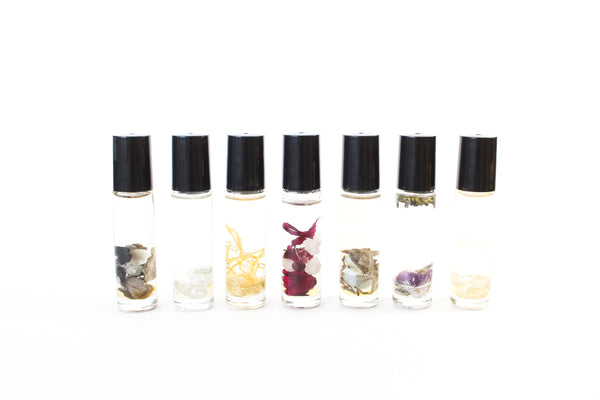 7 clear glass oil roller bottles with black lids, lined up with a white background. They are all filled with different things, based on which chakra they represent. from left to right, sage and black tourmaline, moonstone and silver glitter, calendula and gold glitter, rose and rose quartz with garnet, dried sage and aquamarine,  lavender, amethyst and silver glitter, clear quartz and gold glitter.