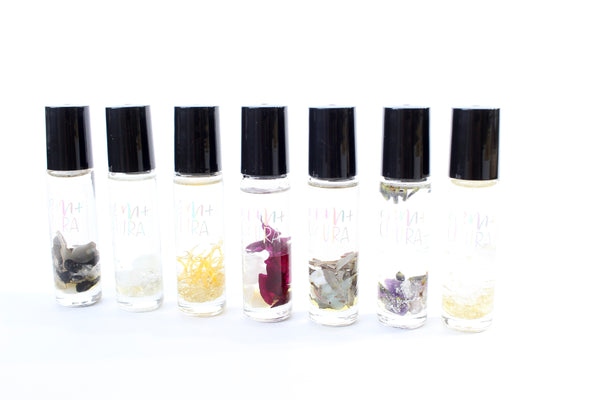 white background with 7 clear glass roller bottles with black lids. the bottles have holographic Gem+Aura logos. in the bottles there is an array of different crystals, dried flowers + herbs. There is one roller for each chakra- displaying the entire roller collection 