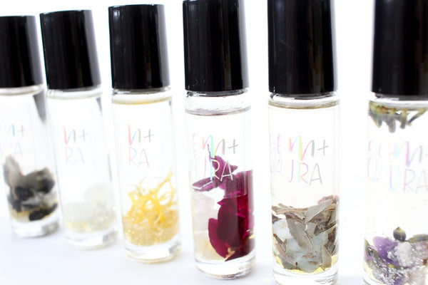 7 clear glass oil roller bottles with black lids, lined up with a white background. They are all filled with different things, based on which chakra they represent. from left to right, sage and black tourmaline, moonstone and silver glitter, calendula and gold glitter, rose and rose quartz with garnet, dried sage and aquamarine, lavender, amethyst and silver glitter, clear quartz and gold glitter. close up photo.