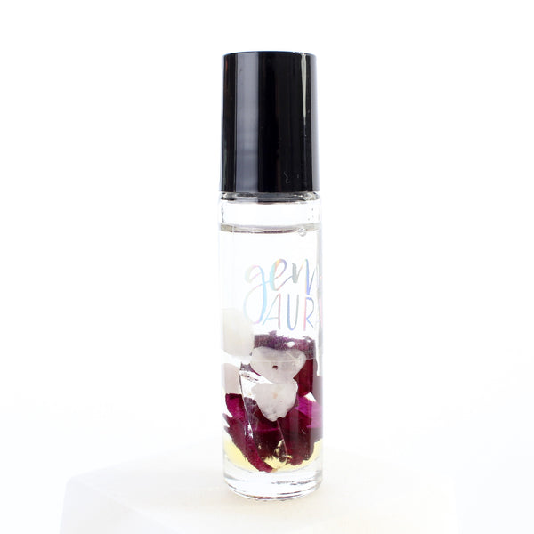 clear glass roller bottle with a black lid and a holographic gem + aura logo on it, in the bottle is an essential oil blend with dried rose petals, rose quartz, garnet and clear quartz inside. there is a white background