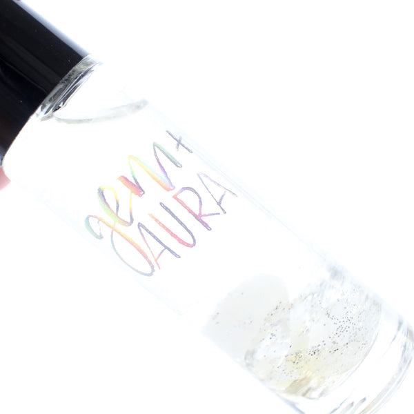 clear glass oil roller bottle with a black lid. It has a holographic Gem + Aura logo on it. The bottle is filled with oil and in it there is clear quartz, moonstone, and mother of pearl with silver glitter. white background. close up.
