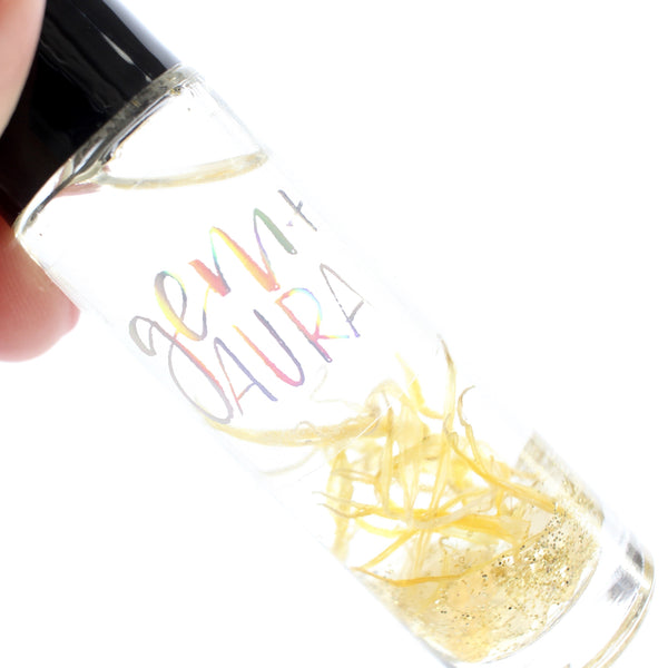 clear glass roller bottle with a black lid and a holographic gem + aura label on it. in the bottle there is an essential oil blend, and dried calendula, gold glitter and citrine chips. there is a white background.