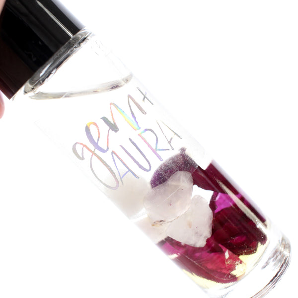 clear glass roller bottle with a black lid and a holographic gem + aura logo on it, in the bottle is an essential oil blend with dried rose petals, rose quartz, garnet and clear quartz inside. there is a white background. close up of contents.