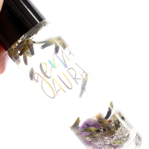 clear glass roller bottle with a black lid and a holographic gem + aura logo on it. bottle is filled with an essential oil blend and also contains silver glitter, dried lavender flowers, and amethyst chips. there is a white background. close up of bottle's contents