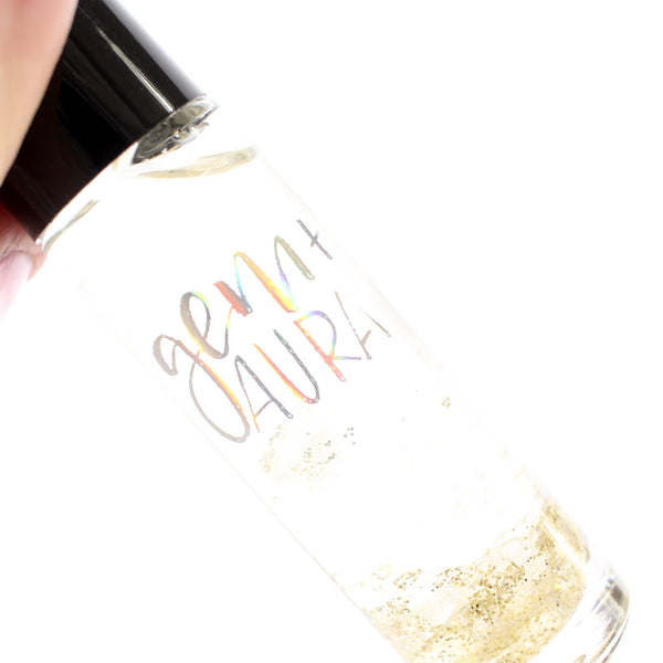 close-up image of white background with a clear glass roller bottle with a black lid. the bottle has a holographic Gem+Aura logo. in the roller is clear quartz and gold glitter